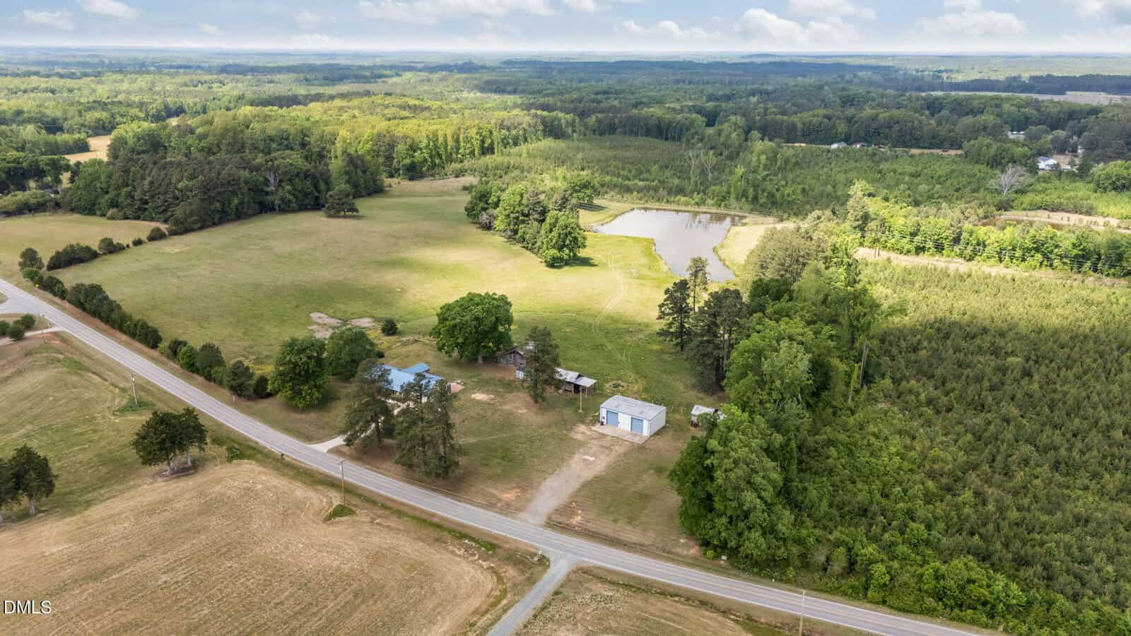 Aerial view of a rural property with open fields, a pond surrounded by trees, several buildings, and a road running along one side, set against a backdrop of forest and distant hills under a partly cloudy sky.