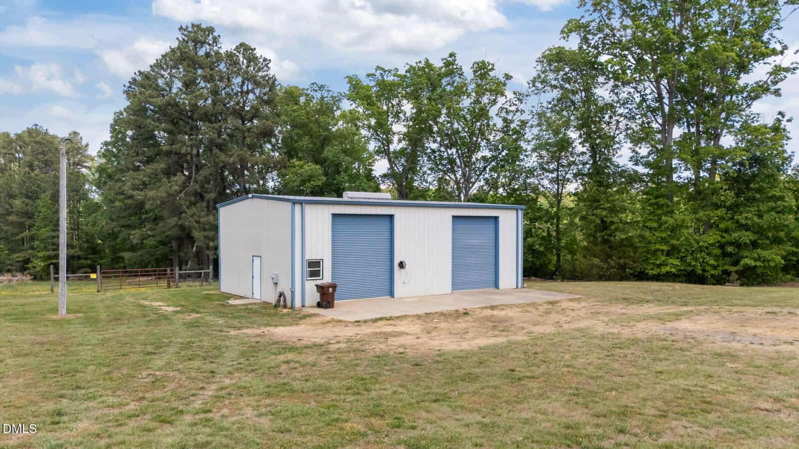 A white metal building with two large blue roll-up doors sits on a concrete pad in a grassy yard, surrounded by trees and a partly cloudy sky overhead.