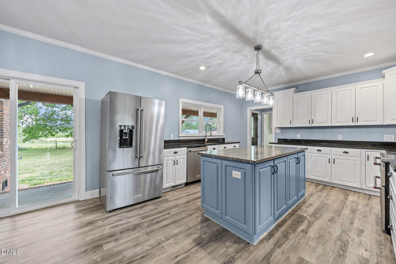 Modern kitchen with light blue walls, white cabinets, stainless steel appliances, a large blue island with a granite countertop, wood-look flooring, and windows showing a green yard outside.