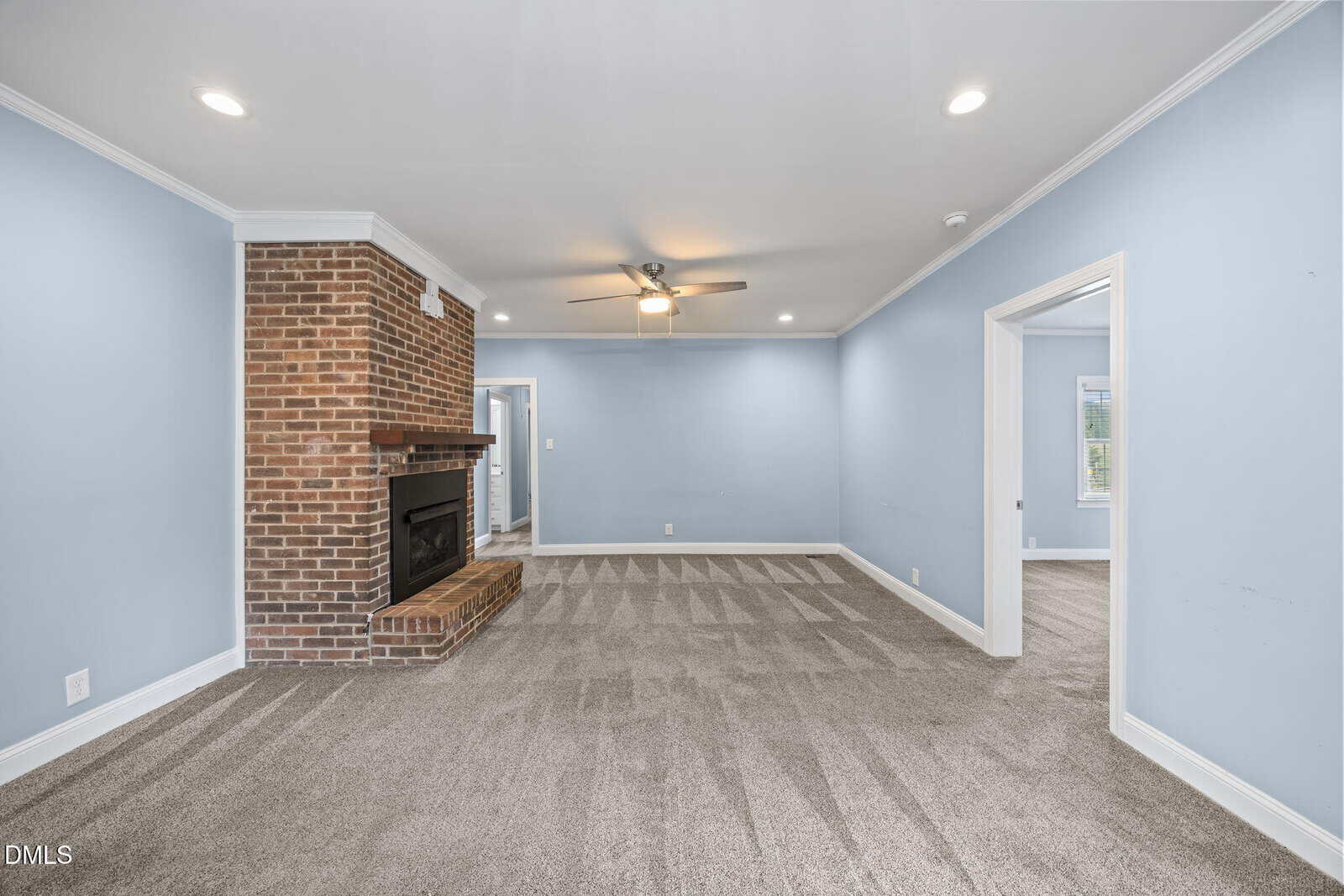 Spacious living room with light blue walls, brick fireplace, ceiling fan, recessed lighting, and gray carpet. Doorways lead to adjacent rooms with natural light.