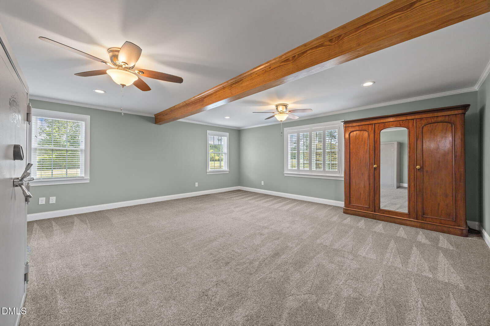 Spacious, carpeted room with light gray walls, wood beam ceiling, two ceiling fans, large windows, and a wooden armoire with mirrored doors. Natural light fills the space.
