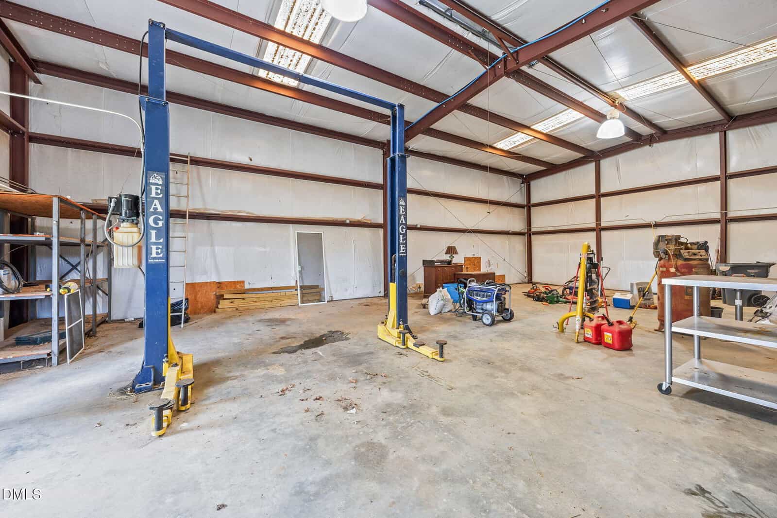 Spacious, well-lit auto repair workshop with a car lift, shelves, tools, and equipment. The concrete floor has some stains, and the space has high ceilings with exposed beams and fluorescent lighting.