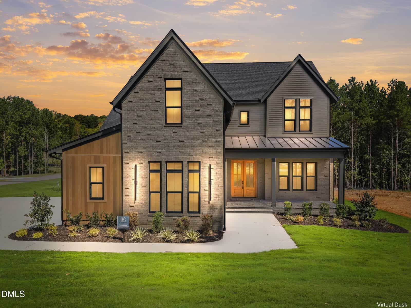 Modern two-story house with brick and siding exterior, large front windows, warm lights illuminating the entrance, manicured landscaping, concrete driveway, and surrounded by trees at sunset—welcome to 150 Cherry Bark Drive in Youngsville.