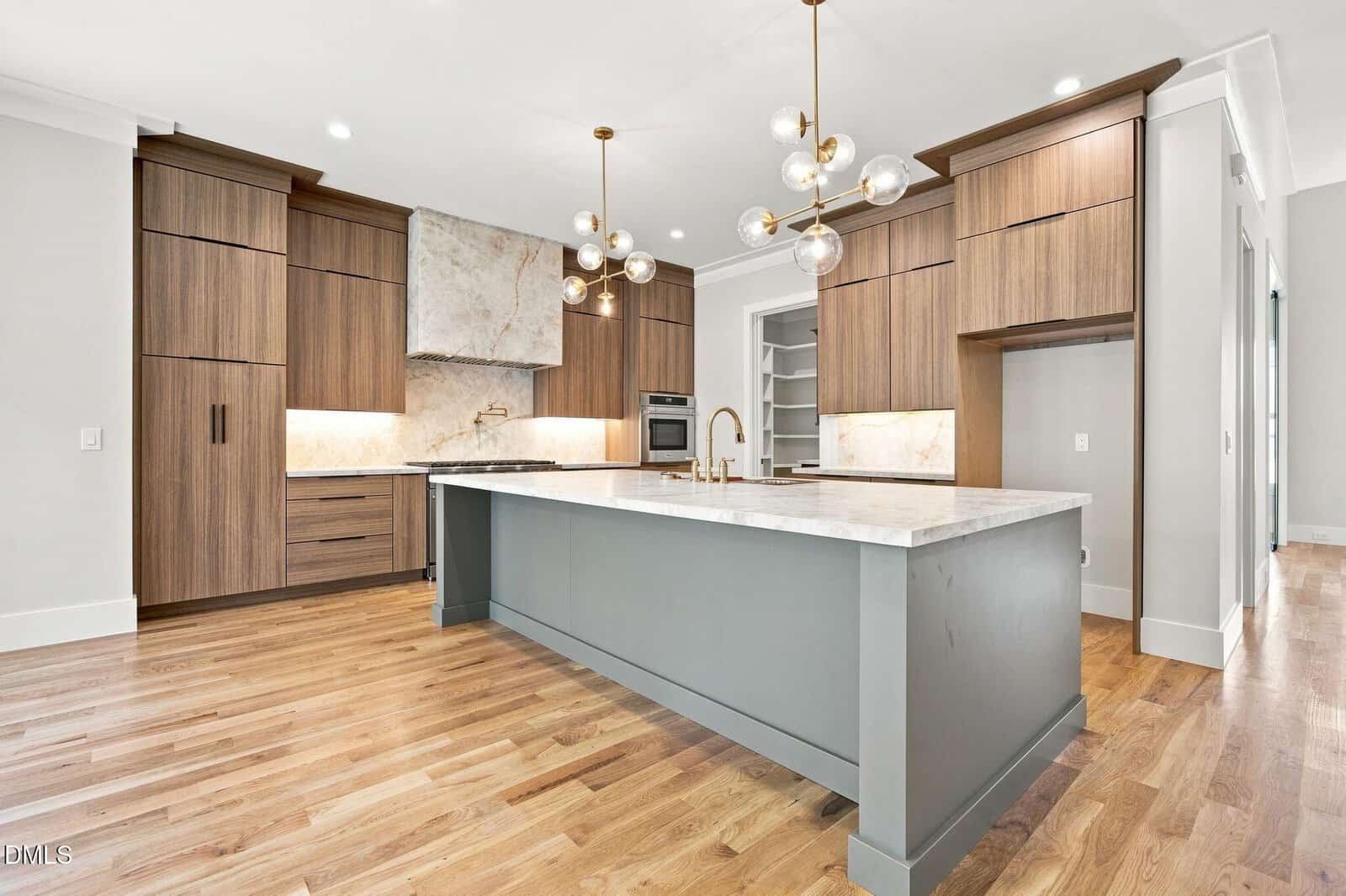 Modern kitchen with wood cabinets, marble countertops, stainless steel appliances, built-in oven, walk-in pantry, light wood floors, and globe pendant lights above a large island.