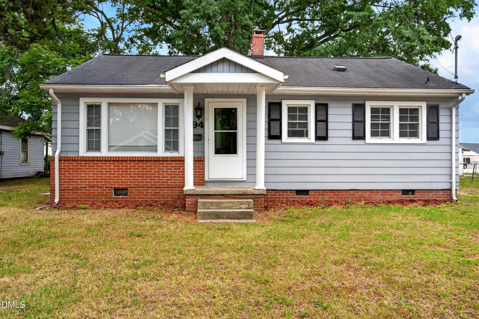 Single-story house with gray siding, red brick foundation, white trim, black shutters, and a covered front porch. The yard has patchy grass, and there are mature trees behind the house.