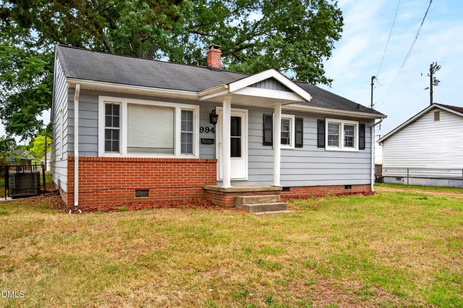 Single-story house with gray siding, black shutters, and red brick foundation. The home features a small covered front porch with white pillars and steps, a large front window, and a grassy yard.
