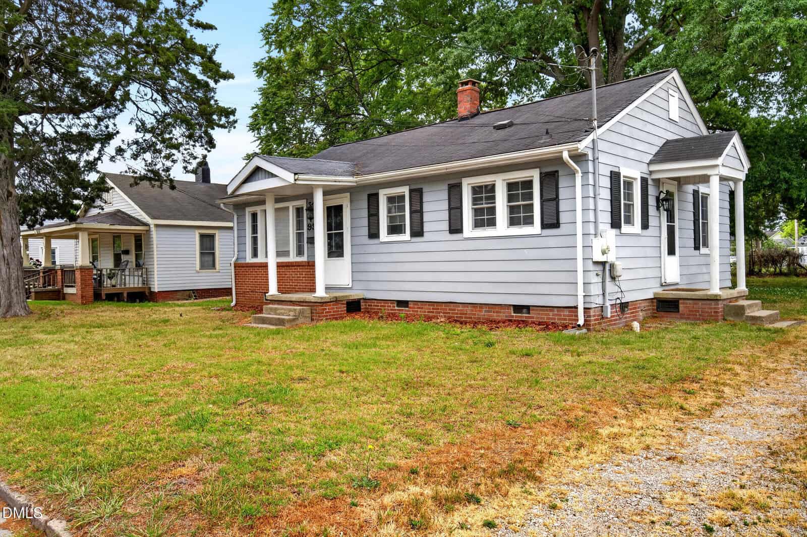 A small, single-story gray house with white trim and black shutters, featuring a brick foundation, front steps, and a grassy yard with some patches of dry grass. Another house is visible to the left.
