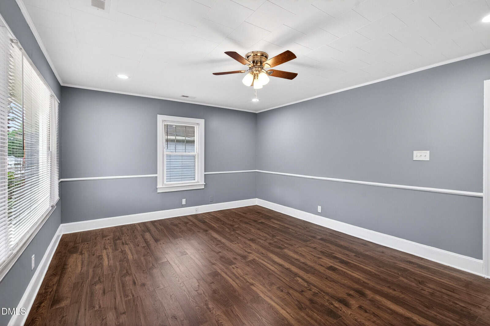A spacious, empty room with gray walls, white trim, dark wood flooring, large windows on one side, and a ceiling fan with lights in the center of the ceiling.