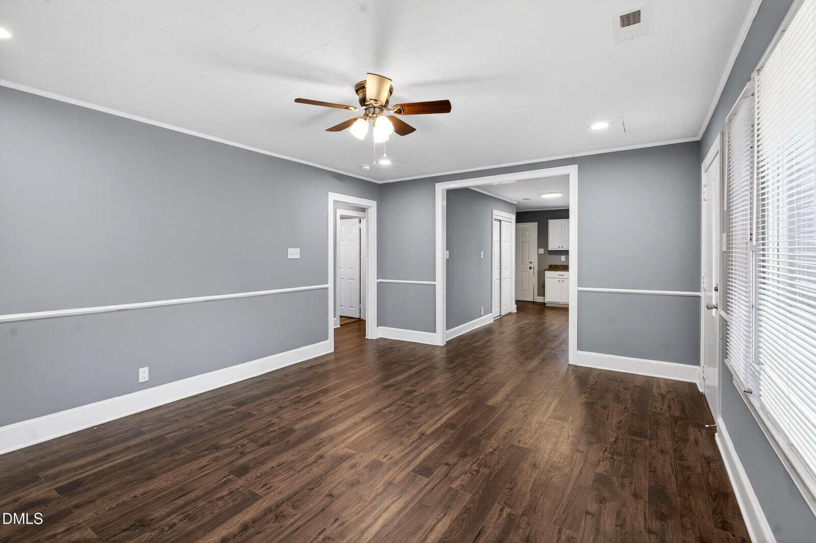A spacious, empty living room with gray walls, white trim, dark wood flooring, a ceiling fan with lights, and large windows. The room opens to a hallway and a kitchen area in the background.