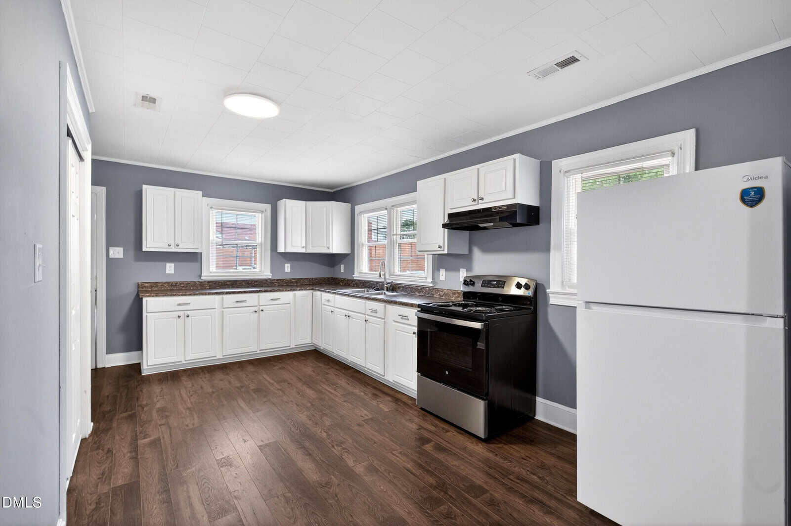 Modern kitchen with dark wood floors, gray walls, white cabinets, a white refrigerator, and a black stove. Three windows provide natural light above the sink and counters. Room appears clean and spacious.
