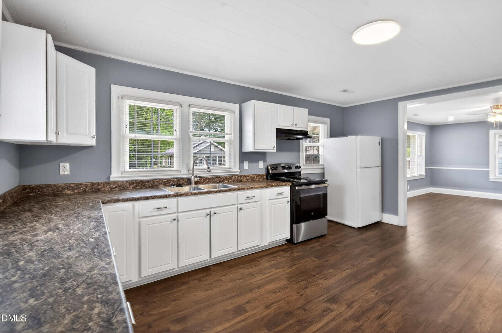 Modern kitchen with white cabinets, dark countertops, stainless steel stove, white fridge, and wood floors. Two windows above the sink and an adjacent dining area with more windows and natural light.