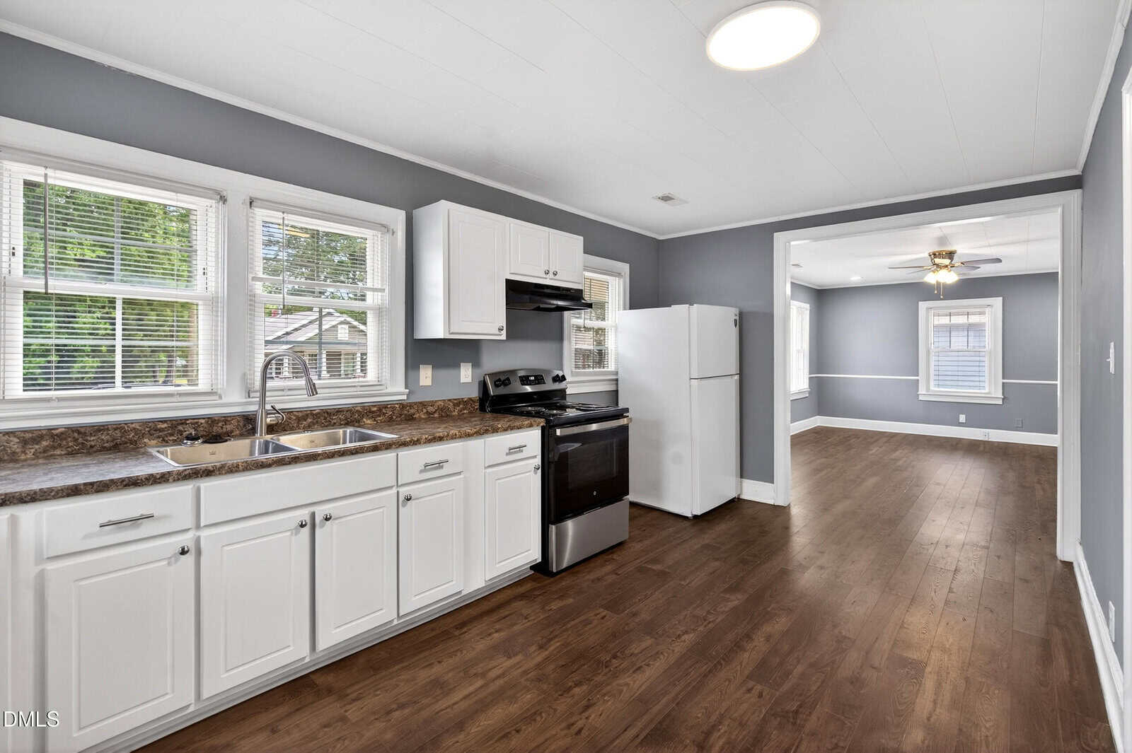 Kitchen with white cabinets, stainless steel sink, stove, and refrigerator. Gray walls, brown countertops, and hardwood flooring. Multiple windows provide natural light; adjacent room with ceiling fan is visible.