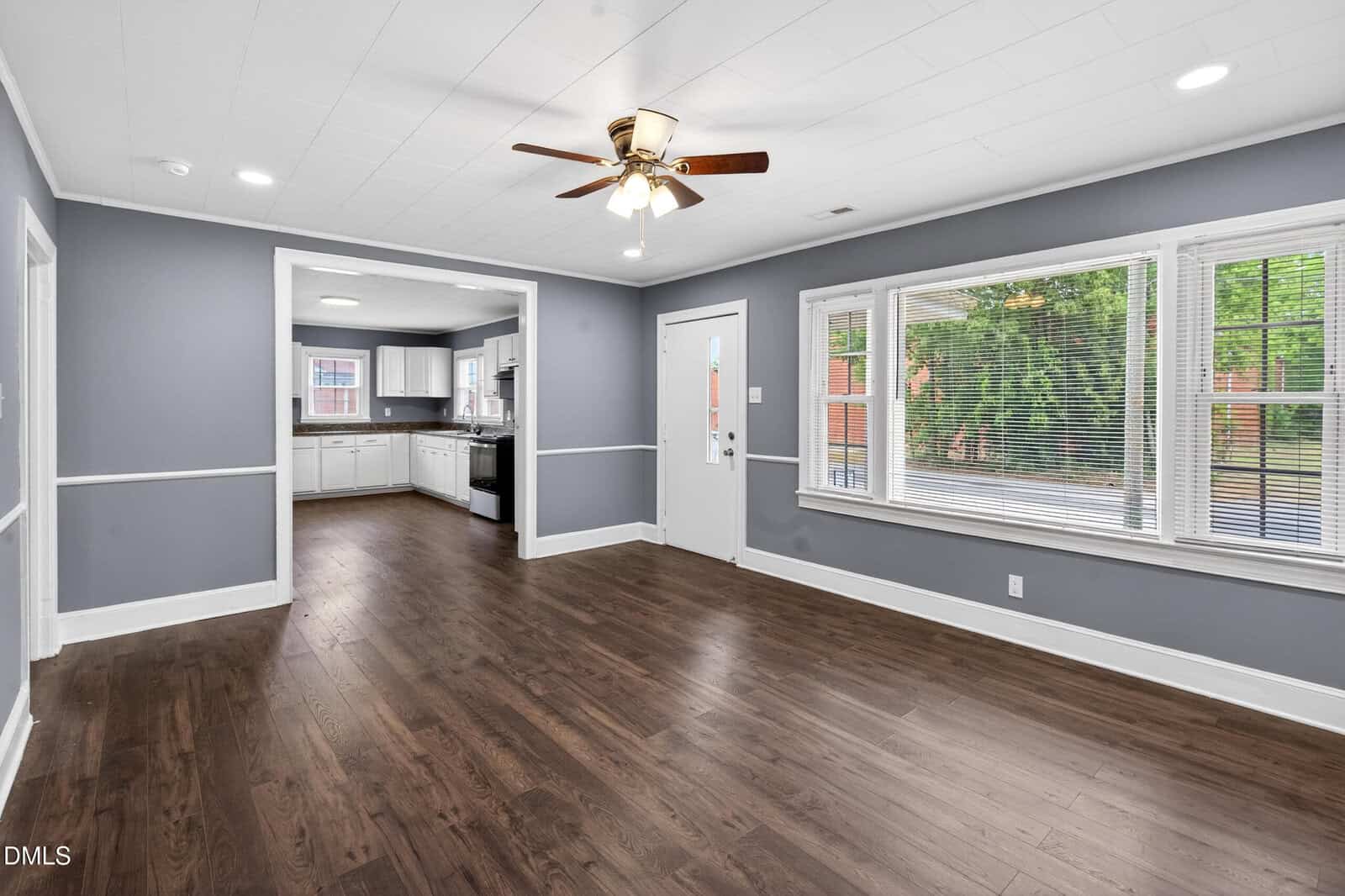 A bright, unfurnished room with dark wood floors, gray walls, white trim, and a ceiling fan. Large windows let in natural light. An open doorway leads to a white kitchen with stainless steel appliances.
