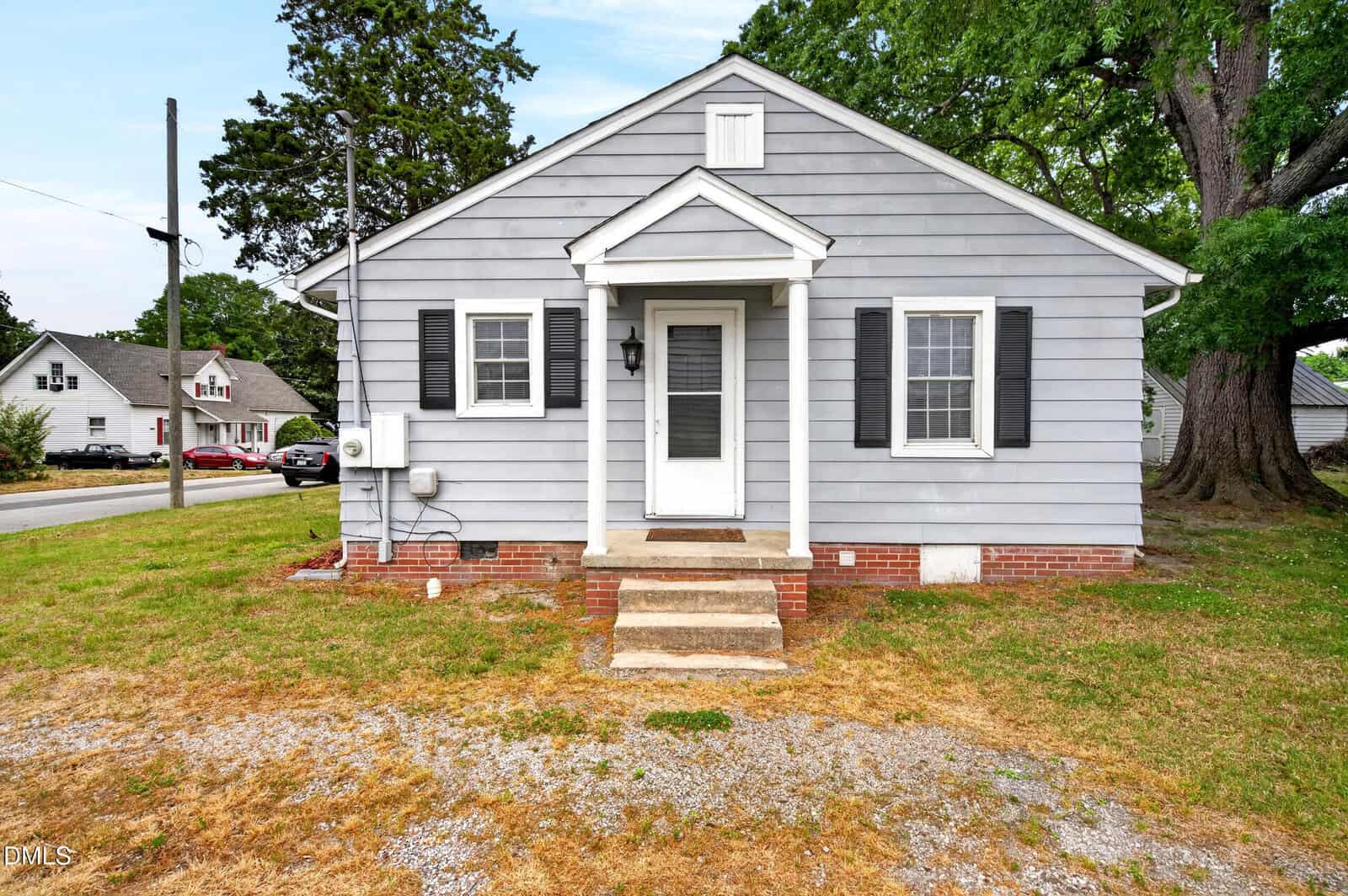 A small, gray single-story house with white trim, black shutters, and a covered front porch. The yard has patchy grass, and nearby trees and houses are visible in the background.