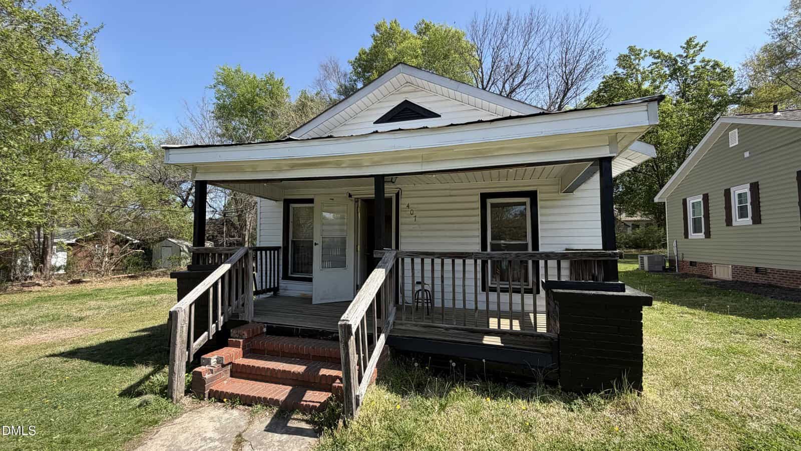 A small, white, single-story house with a covered front porch, black trim, and steps leading up to the entrance. The yard is grassy with trees in the background and a neighboring house to the right.