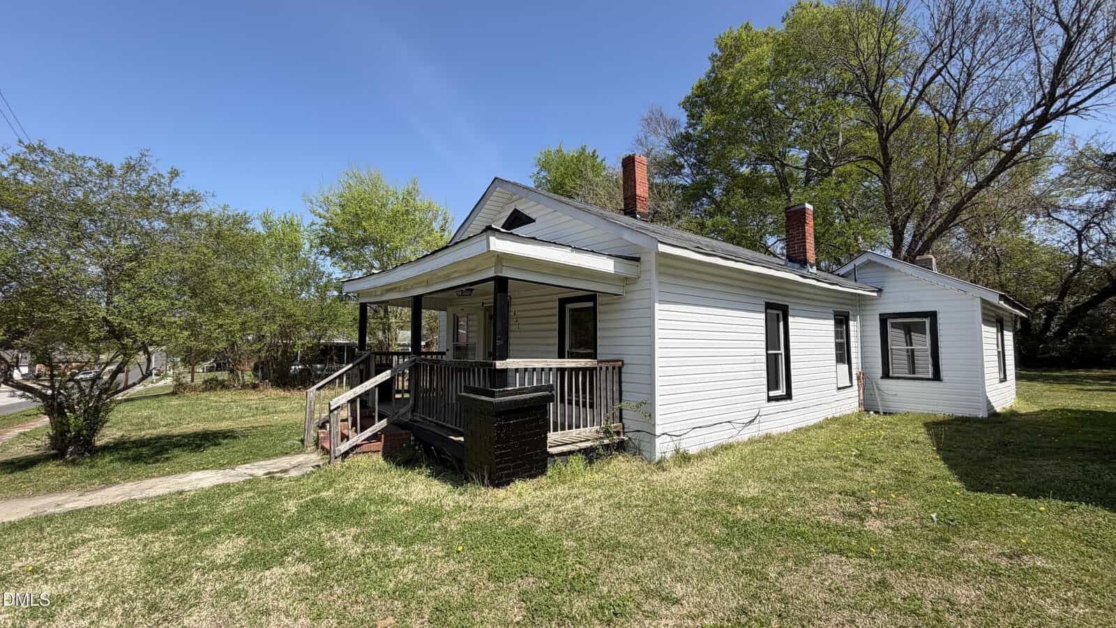 A small, white, single-story house with a covered front porch, two brick chimneys, and black trim sits on a grassy lot with trees in the background under a clear blue sky.