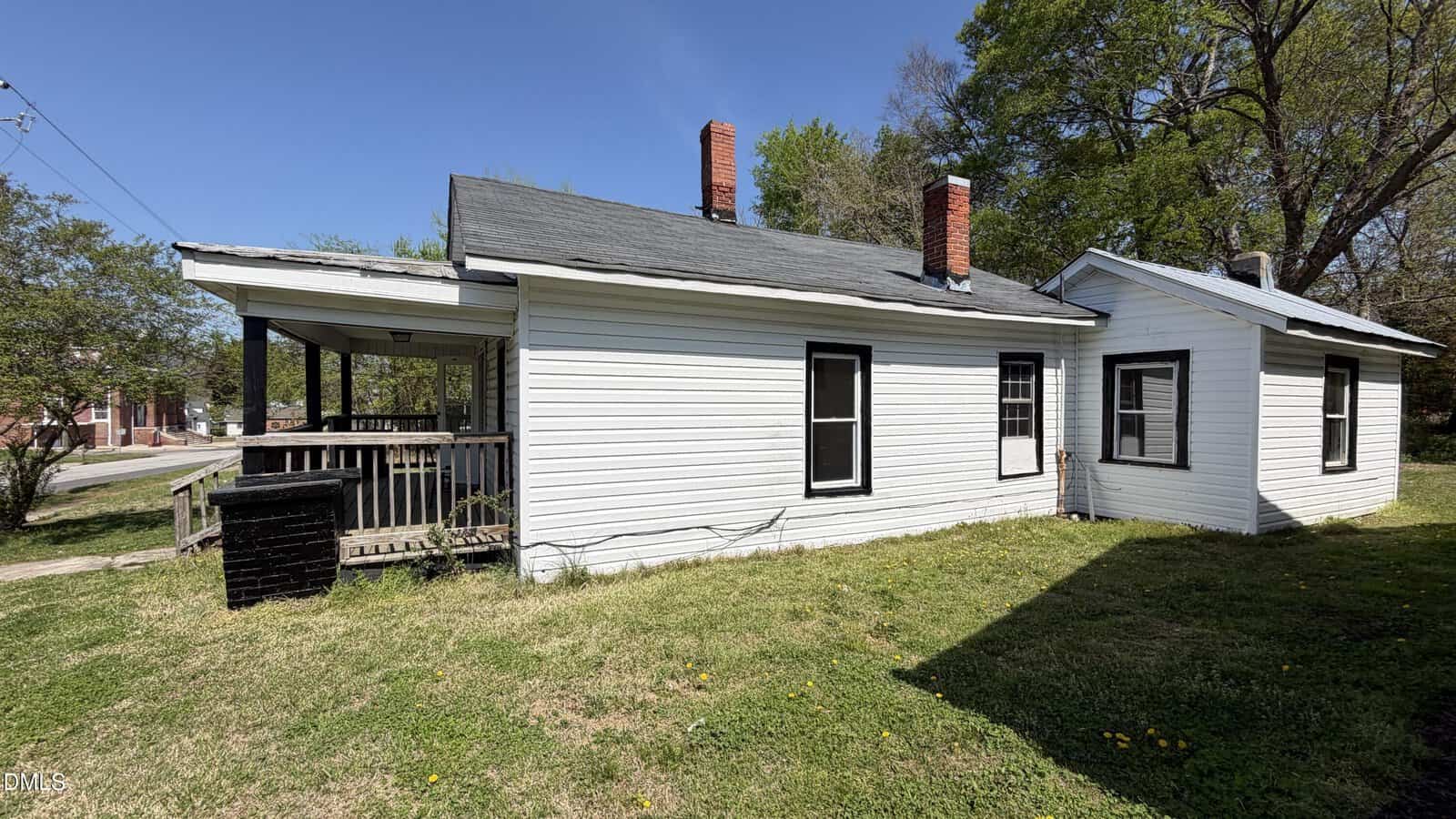 A small white house with black trim and two red brick chimneys sits on a grassy yard under a clear blue sky. A covered porch is visible on the left, and large trees are in the background.