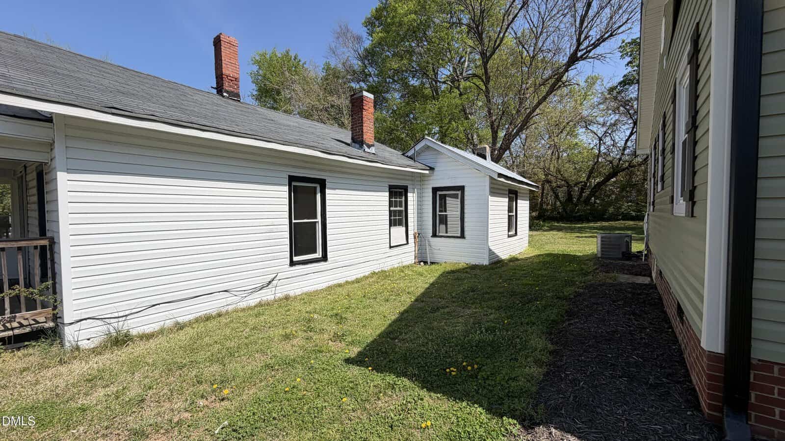 A white, single-story house with black-trimmed windows and two red brick chimneys sits beside a green lawn on a sunny day. Another building with white siding and green trim is partially visible on the right.