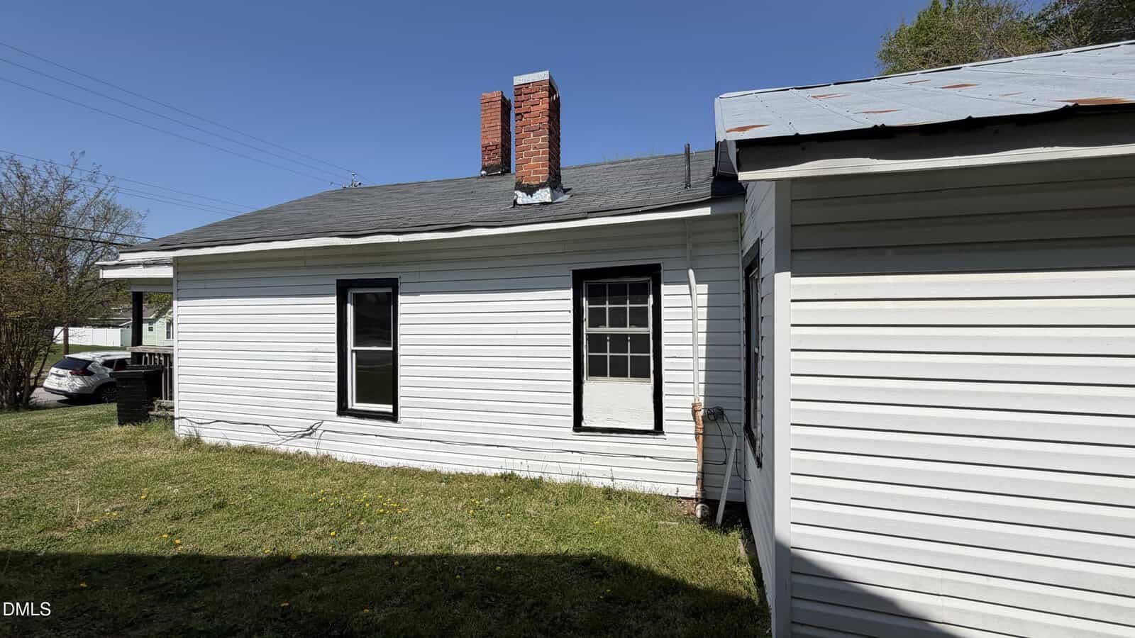 A white house with black-trimmed windows, a grassy yard, a brick chimney, and a small attached structure with a metal roof under a clear blue sky.