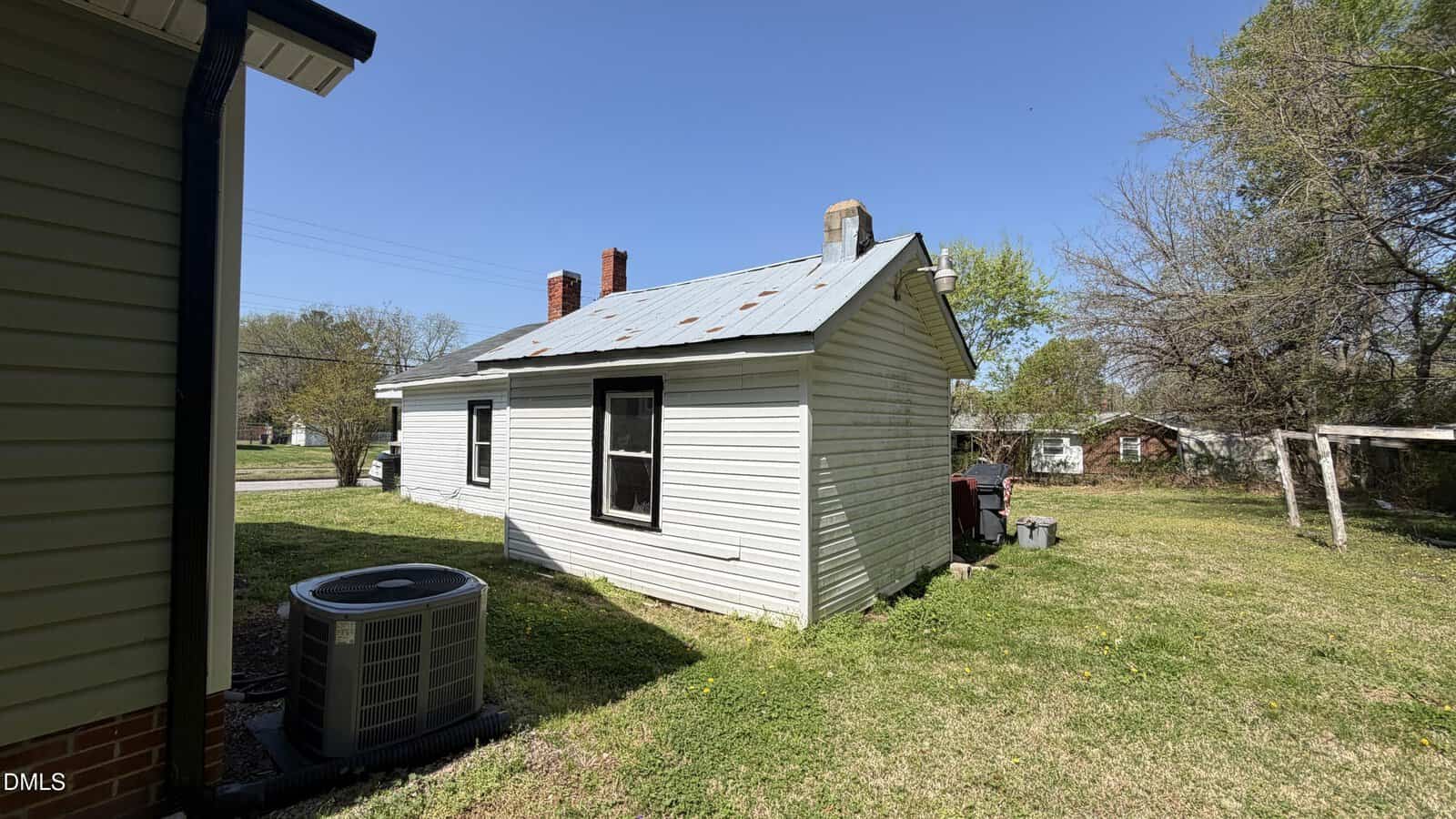 A small, white house with a metal roof stands on a grassy yard. An air conditioning unit is visible in the foreground, with trees and other homes in the background under a clear blue sky.