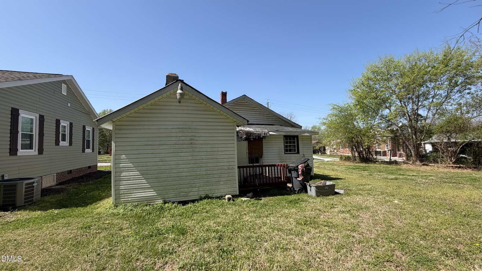 Small white house with a covered porch and weathered roof, surrounded by grass and trees under a clear blue sky. Adjacent houses and yard equipment are visible.