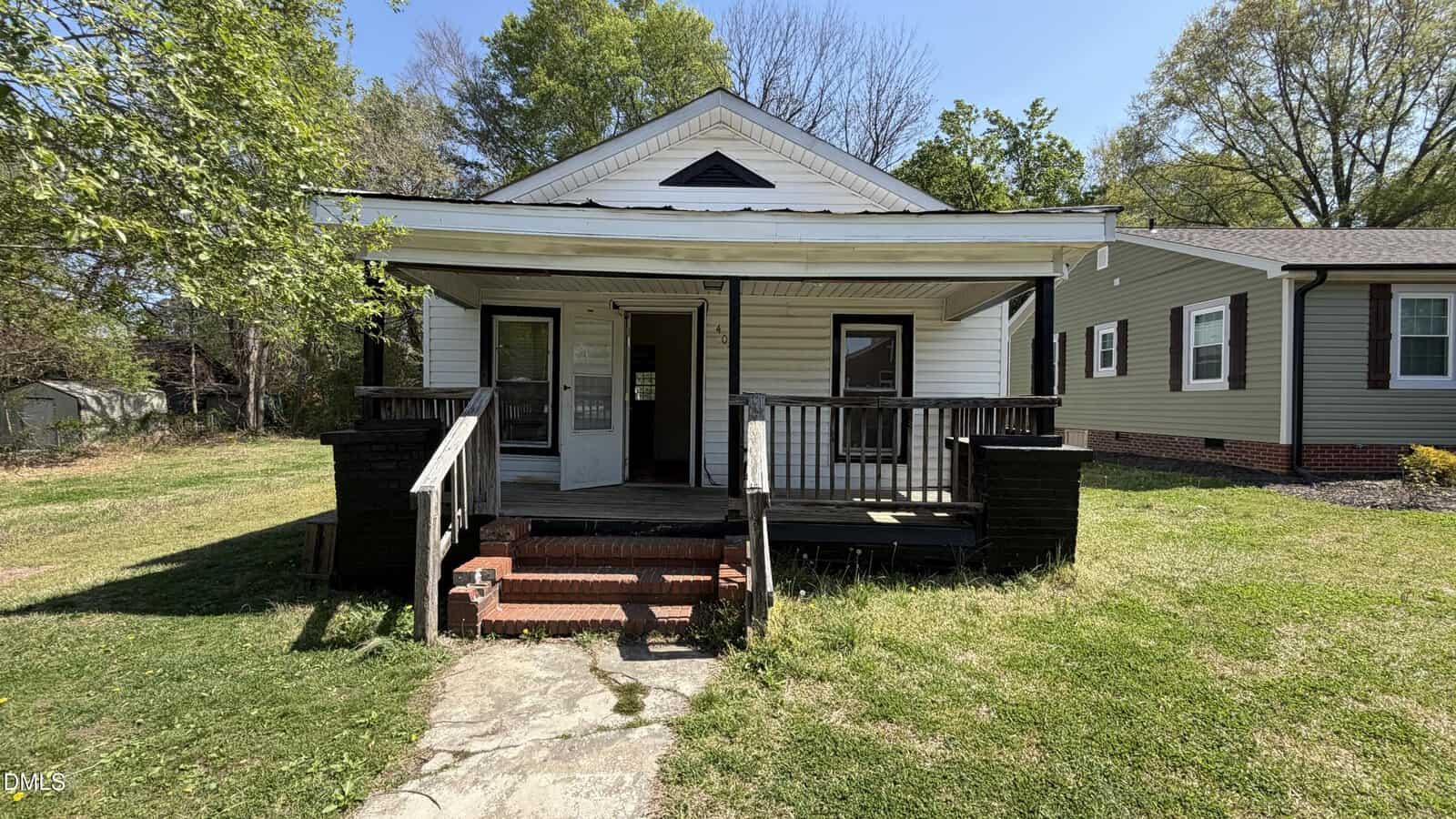 A small, white, single-story house with a covered front porch, black railings, and steps leading to the entrance. The yard is grassy with trees and another house visible to the right.