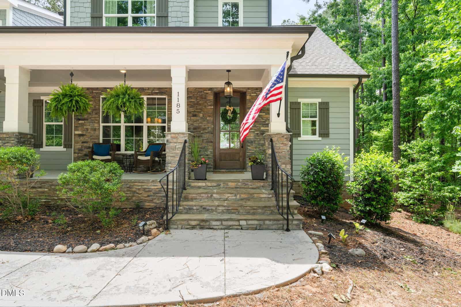 A stone-front porch with steps, black railings, two hanging ferns, an American flag, seating area with blue cushions, and a wooden door, surrounded by green shrubs and trees. House number 1185 is visible by the door.