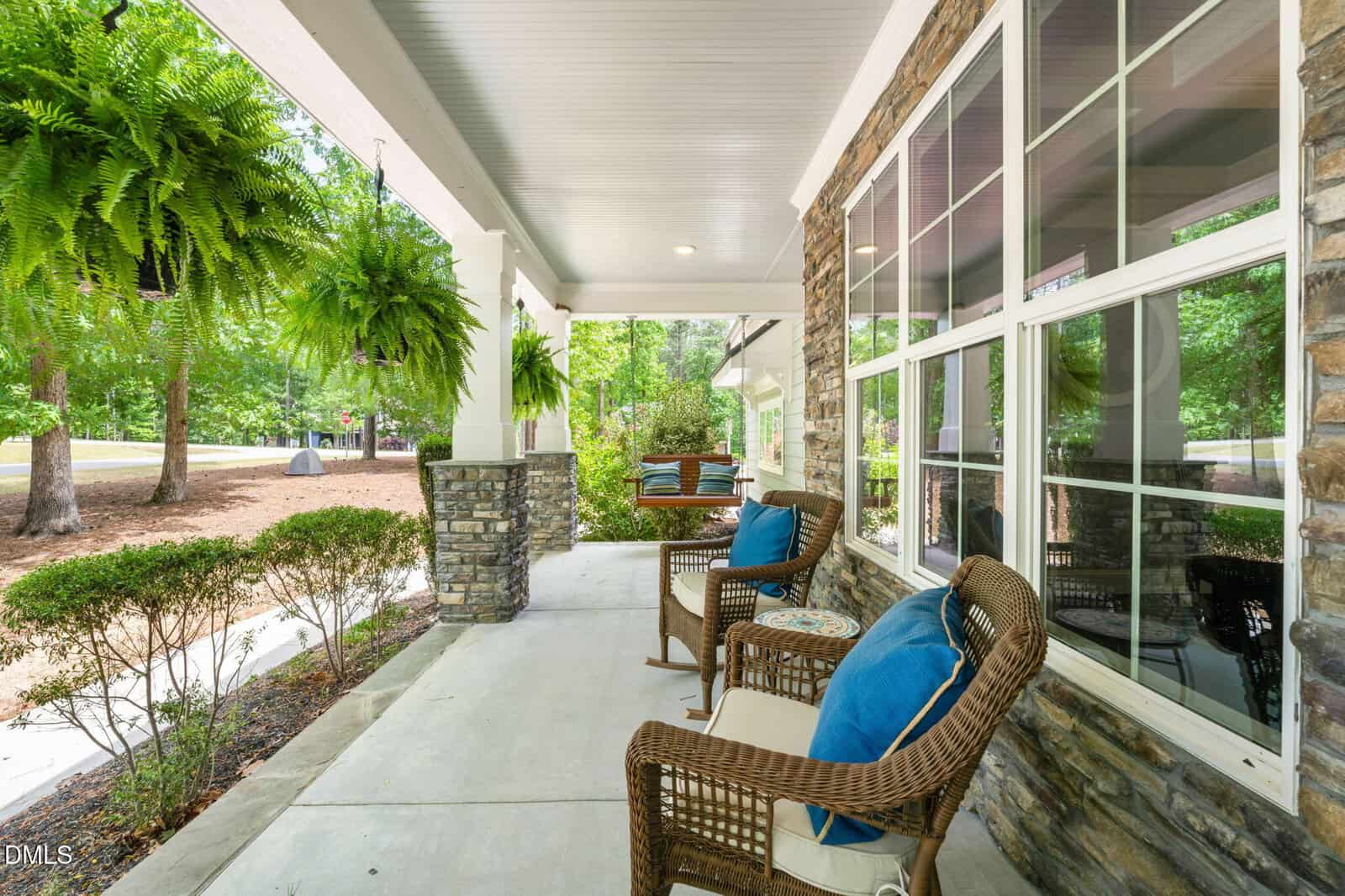 A cozy front porch with wicker chairs, blue cushions, hanging ferns, and stone columns overlooking a yard with trees and shrubs on a sunny day.