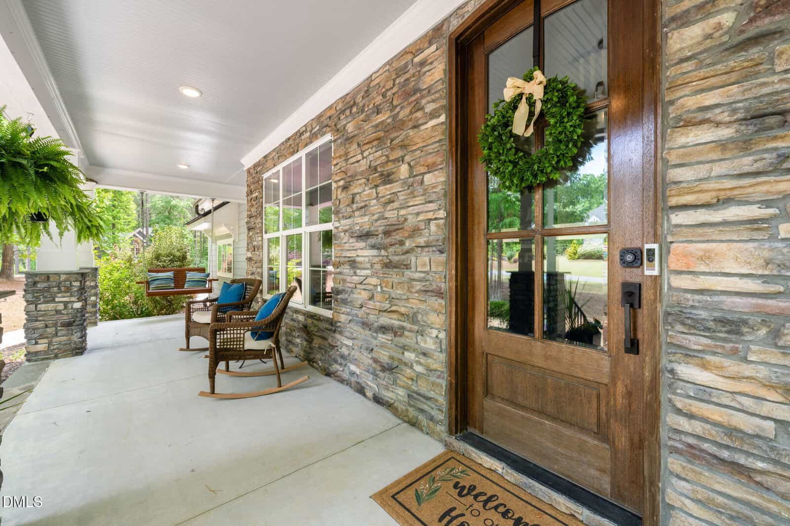 A front porch with stone walls, a wooden door with a wreath, welcome mat, rocking chairs with cushions, a hanging fern, and a swing bench. Trees and greenery are visible in the background.