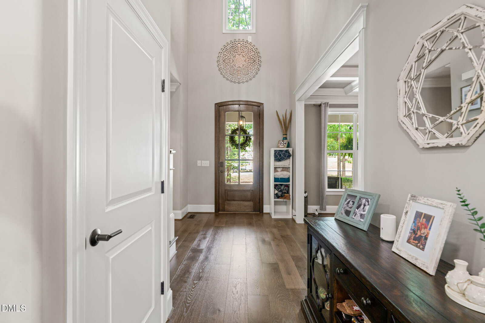 Bright, airy entryway with wood flooring, tall ceiling, a wooden front door with a wreath, decorative shelves, and a console table displaying framed photos and decor, with sunlight streaming through windows.
