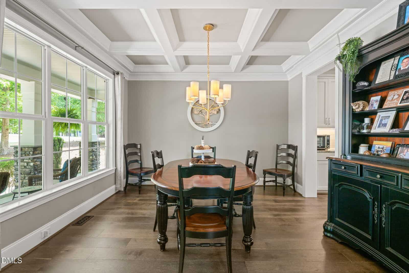 Bright dining room with a wooden table, six chairs, a chandelier, large window, decorative wall clock, and shelving unit displaying photos and plants. Coffered ceiling and light color scheme enhance the elegant space.