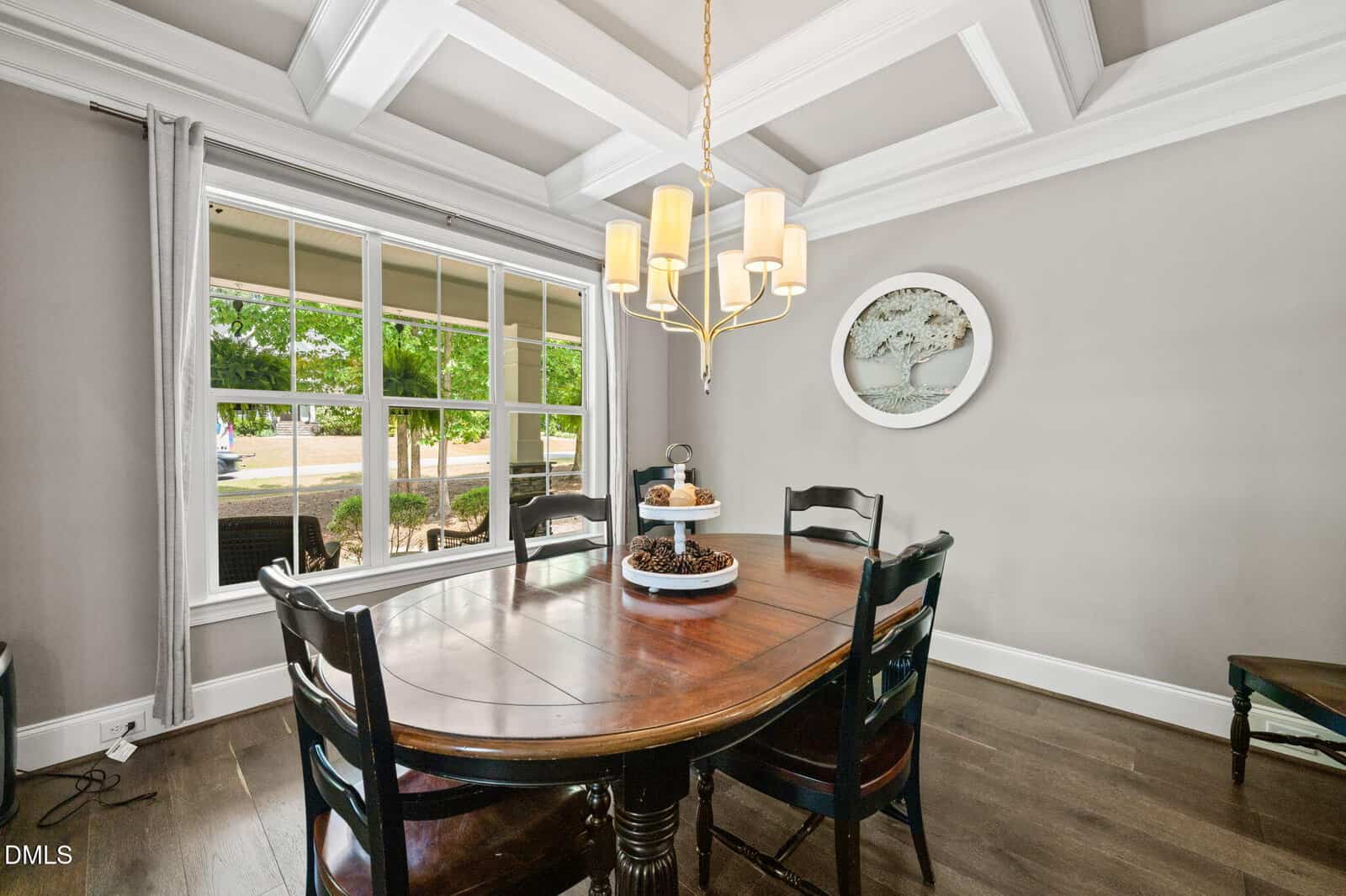 Bright dining room with a dark wooden table, four chairs, a tiered tray centerpiece, large window with gray curtains, coffered ceiling, and a round tree art piece on the gray wall.