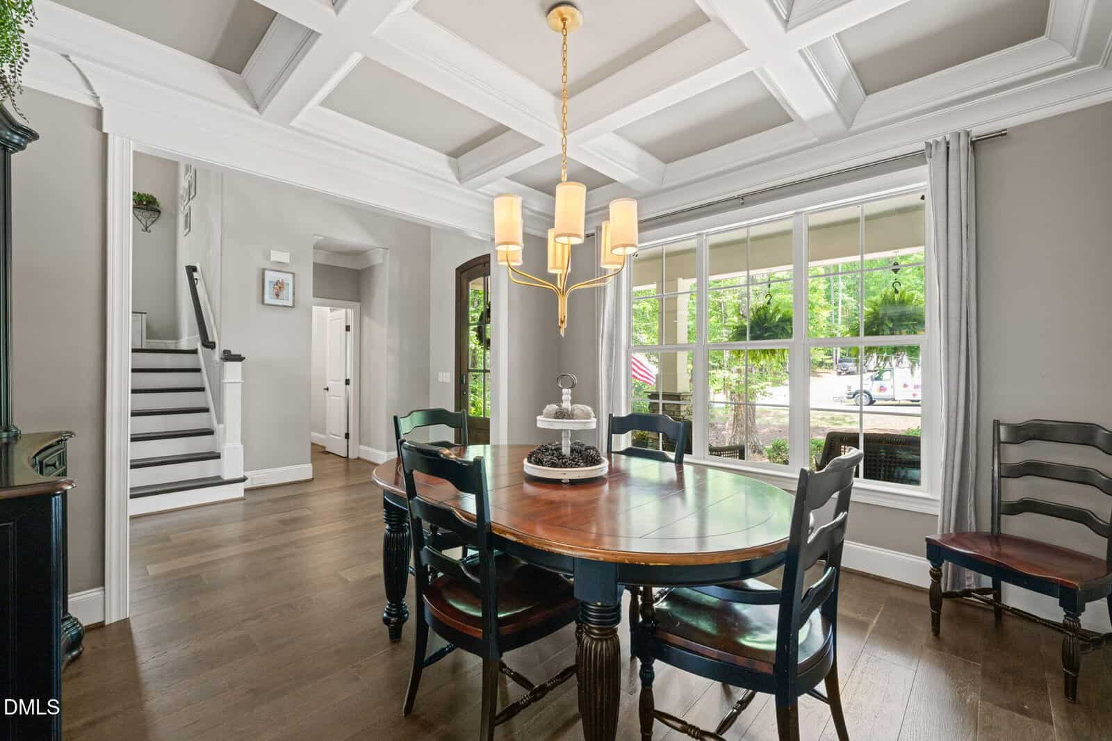 A dining room with a round wooden table, five chairs, a chandelier, large window with curtains, and coffered ceiling. A staircase and hallway are visible in the background.