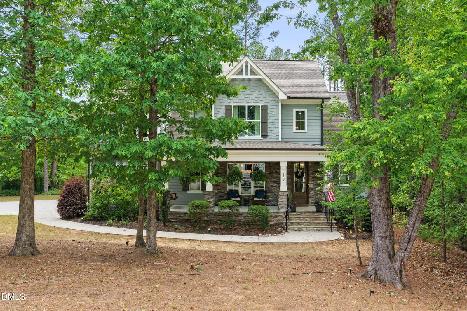 A two-story house with gray siding and white trim, a front porch with chairs, and surrounded by tall trees and a wooded yard. An American flag hangs near the porch, and a curved driveway leads to the home.