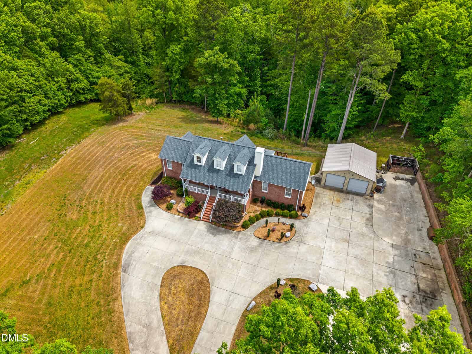 Aerial view of a two-story brick house with a gray roof, front porch, and dormer windows, surrounded by trees. There's a large driveway, landscaped yard, and a separate garage building to the right.