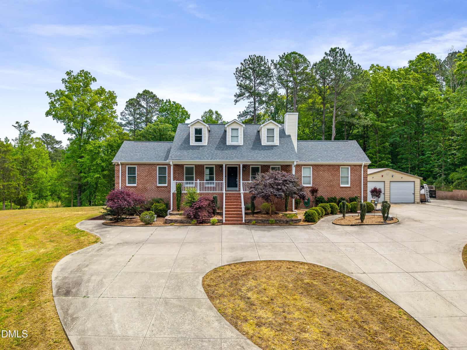 A brick house with dormer windows and a covered front porch sits on a large lot with a circular driveway, landscaped shrubs, and a detached garage surrounded by trees and greenery.