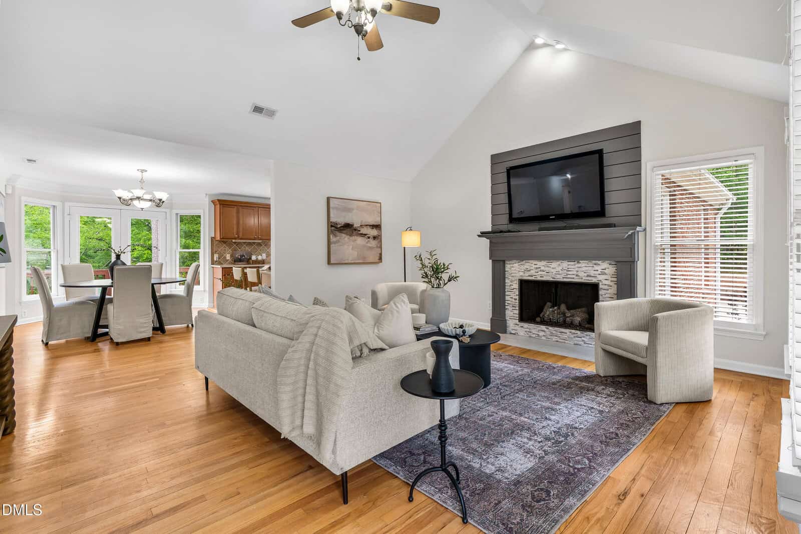 Bright living room with light-colored sofas, armchair, a dark area rug, and a fireplace with a mounted TV above. Hardwood floors, large windows, and an open view into the dining area and kitchen create an airy, welcoming space.