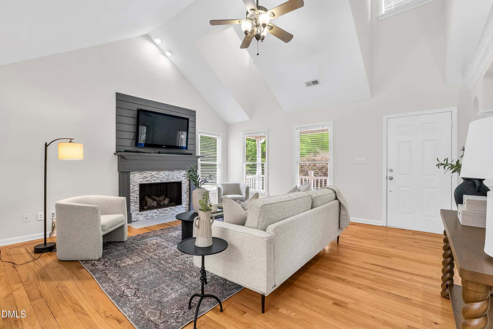 Bright living room with a ceiling fan, fireplace, mounted TV, light sofas, round table, armchair, floor lamp, and hardwood floors. Large windows provide natural light, and a rug adds warmth to the cozy space.