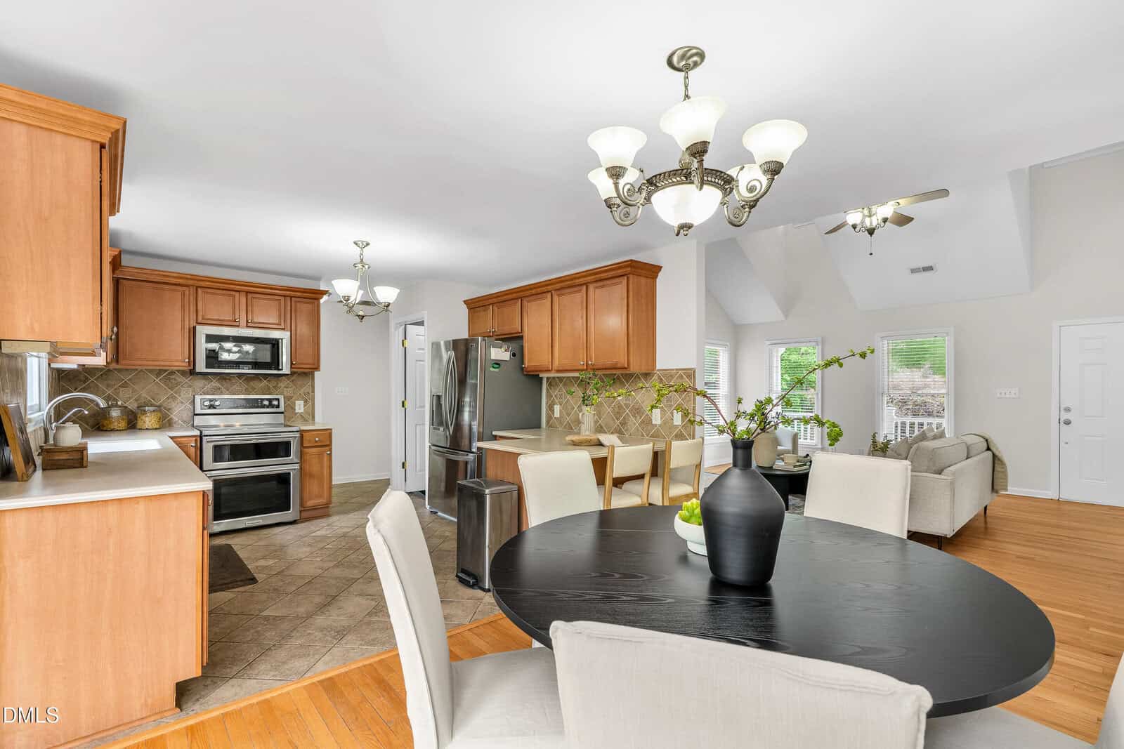 Bright, modern kitchen and dining area with wooden cabinets, stainless steel appliances, round black dining table with beige chairs, and a cozy living space in the background. Multiple chandeliers hang from the ceiling.