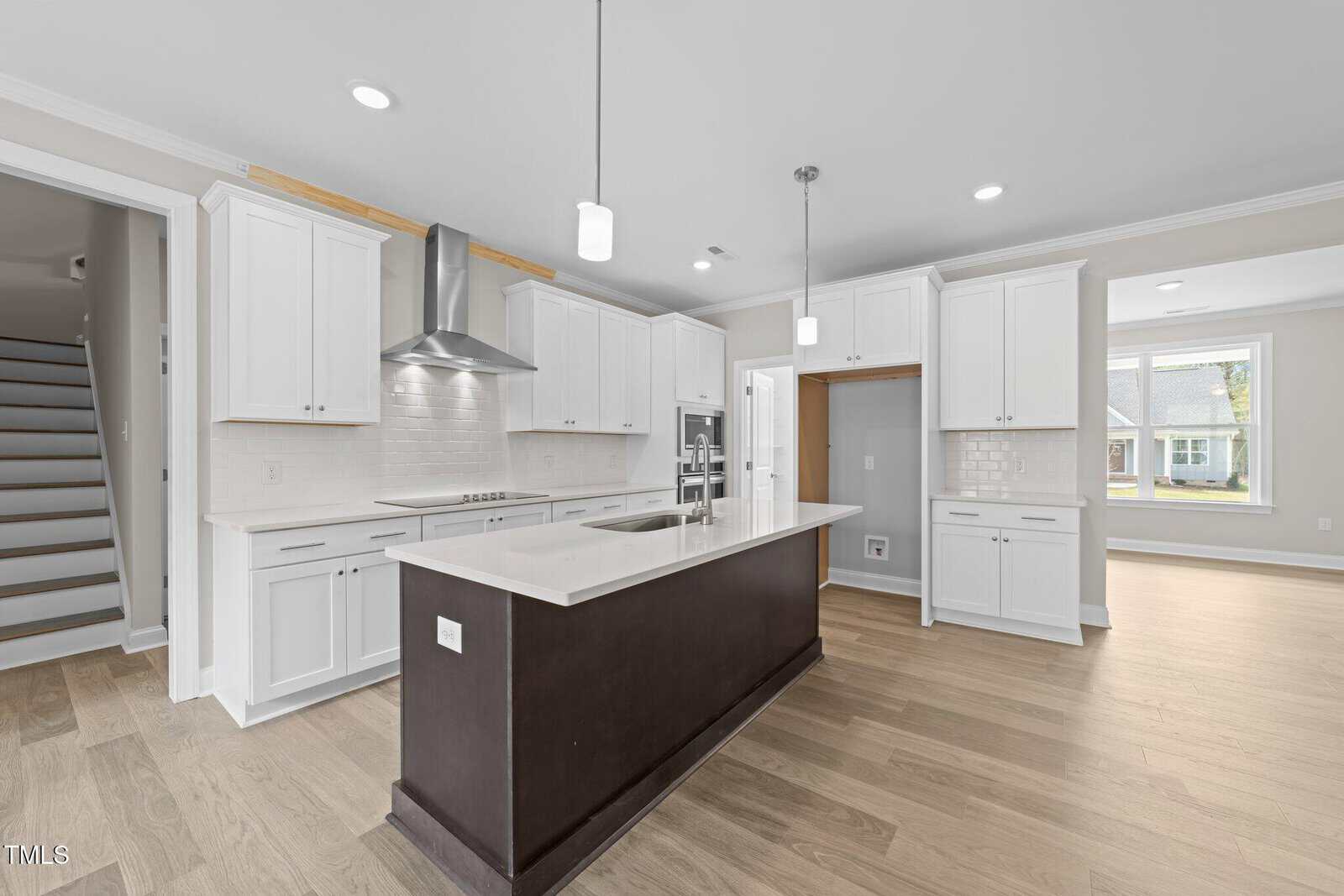 Modern kitchen with white cabinets, stainless steel appliances, a dark island with a sink, light wood flooring, pendant lighting, and large windows letting in natural light. A staircase is visible on the left.