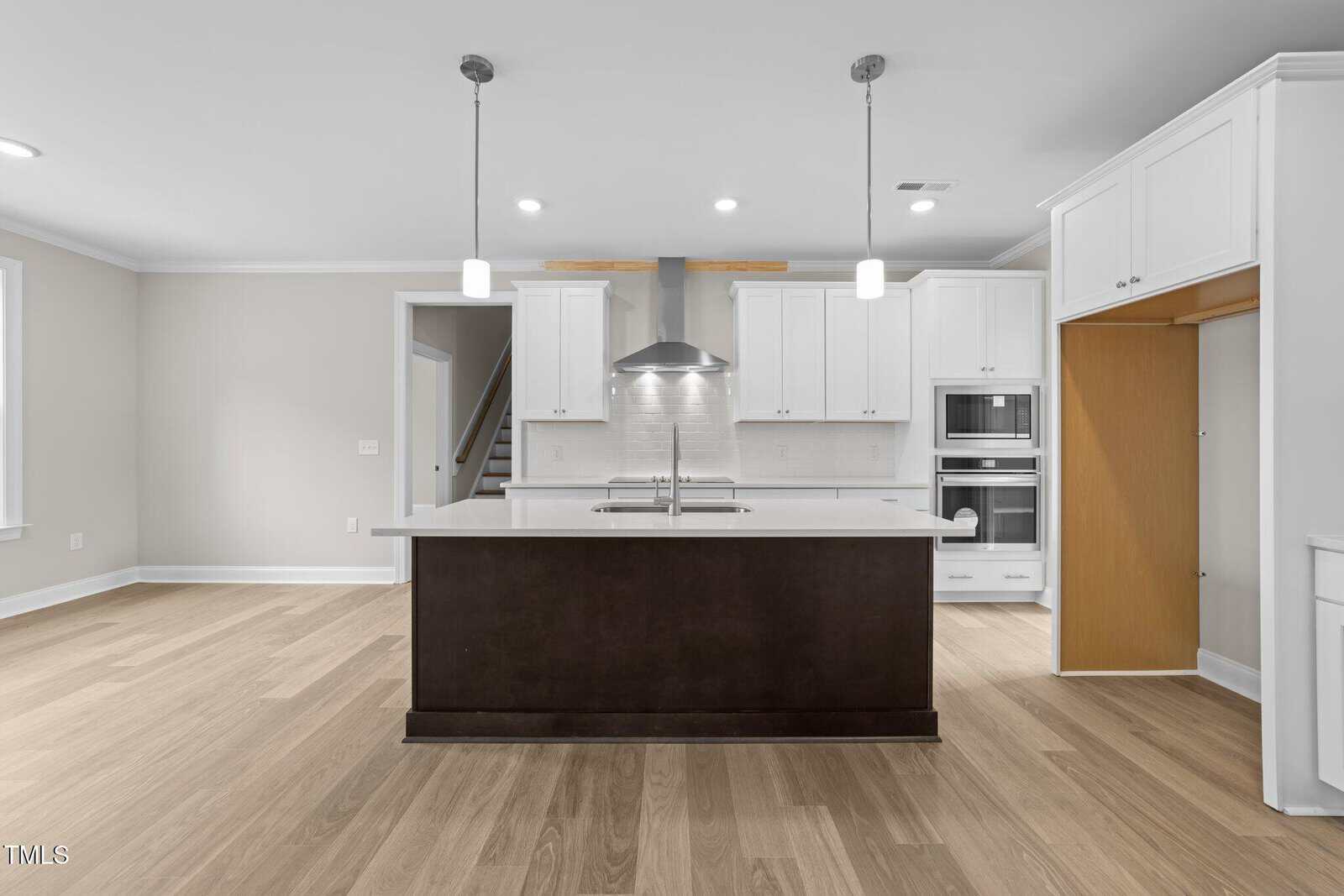 Modern kitchen with a dark island, white cabinets, built-in ovens, and stainless steel range hood. Light wood flooring and pendant lights, with an open pantry and visible staircase in the background.