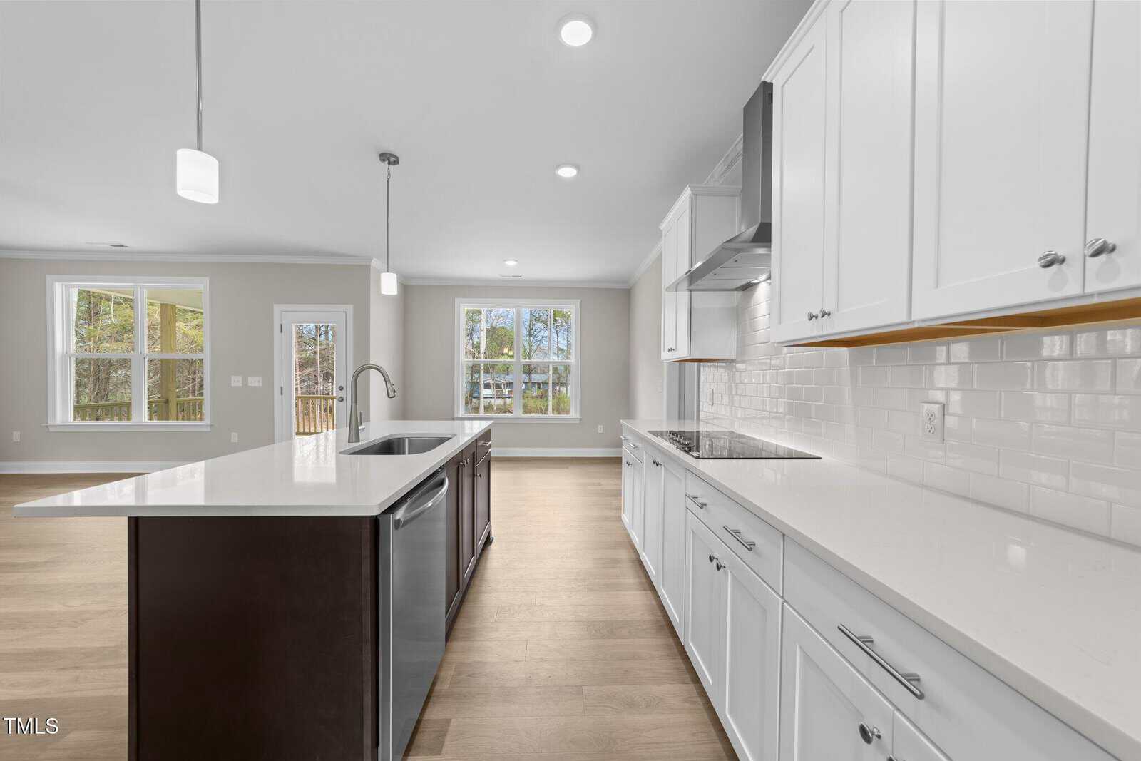 Modern kitchen with white cabinets, stainless steel appliances, an island with dark wood base, and pendant lights. Large windows let in natural light. Light wood flooring throughout. Open layout connects to living area.