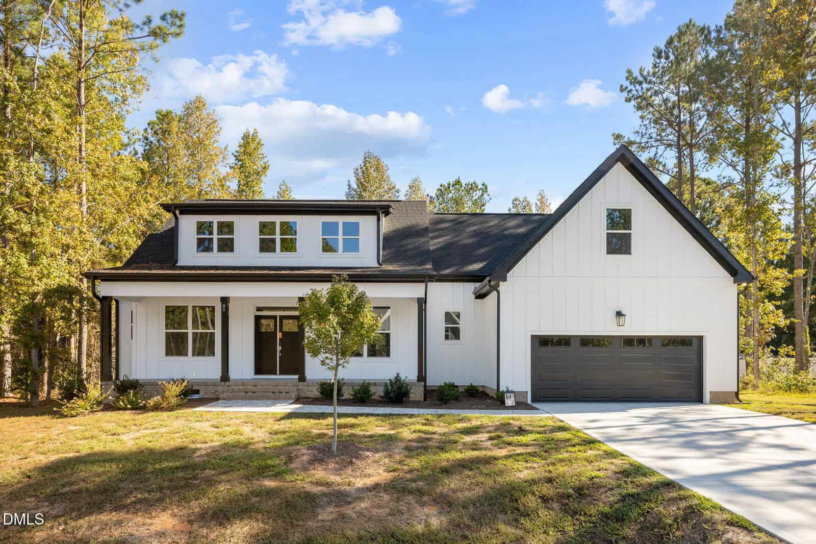A modern white farmhouse with black accents, a covered front porch, and a two-car garage is surrounded by trees and sits under a clear blue sky.