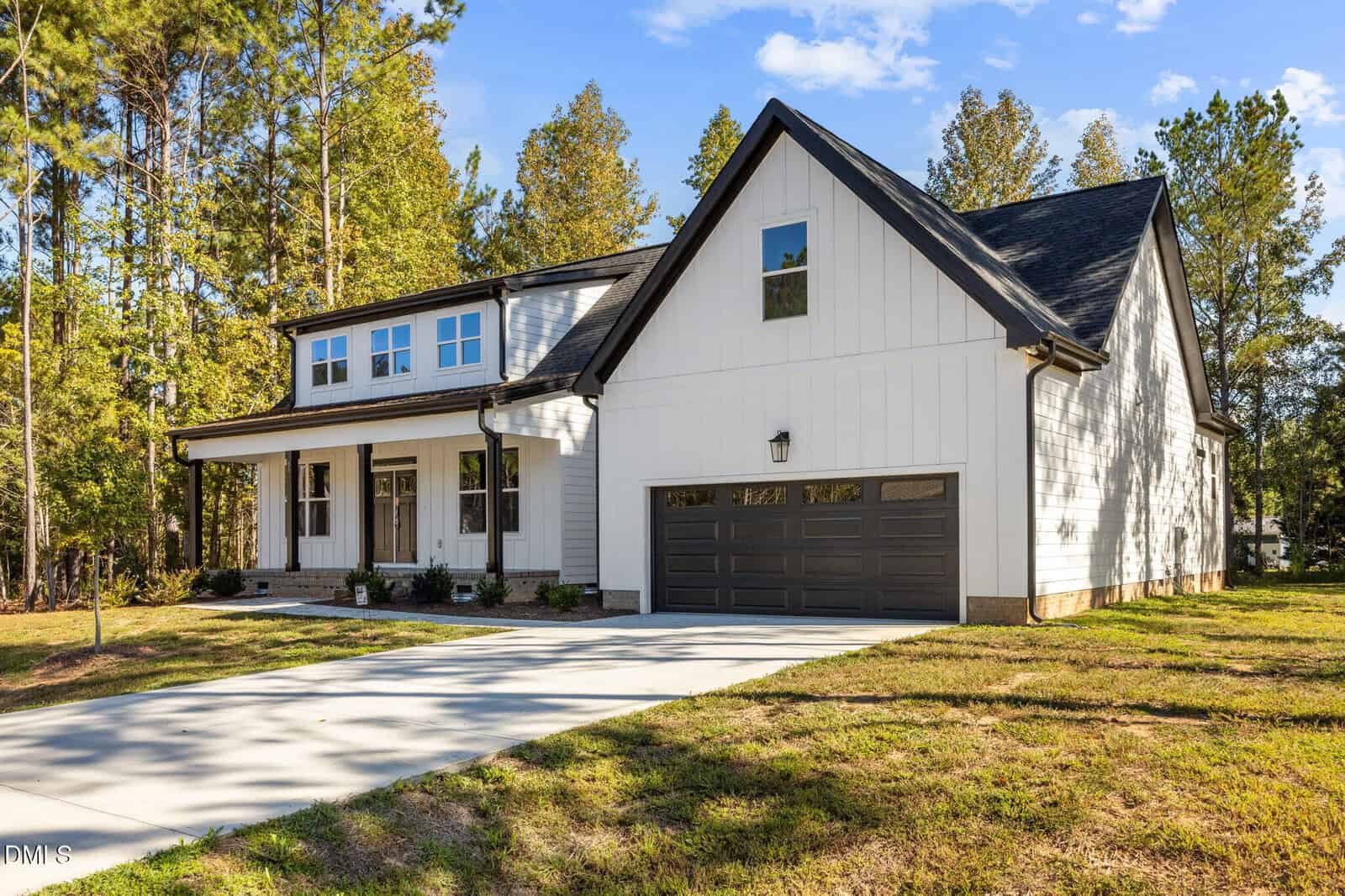 A modern two-story white house with dark trim, a large front porch, and a double garage sits in a grassy yard surrounded by tall trees under a blue sky.