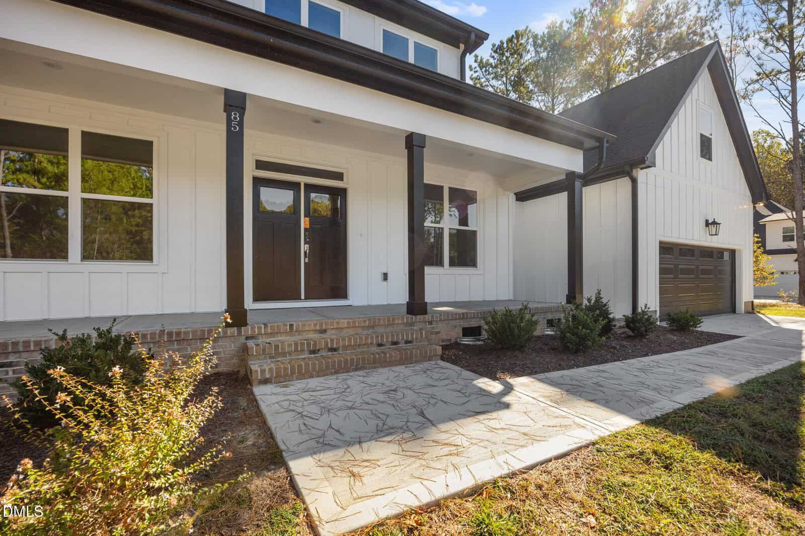 A modern white house with large windows, a dark front door, a covered front porch with brick steps, landscaped shrubs, a concrete walkway, and an attached two-car garage.
