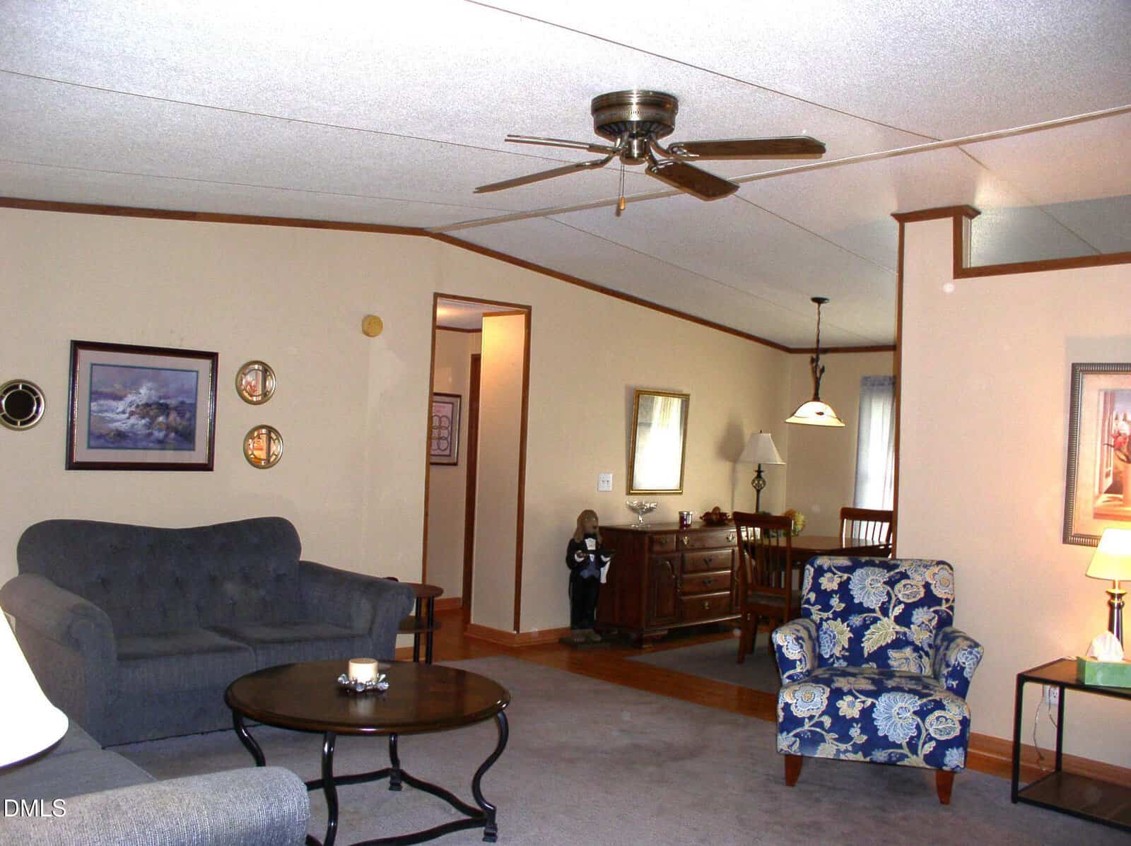 A cozy living room with beige walls, a gray sofa, a round coffee table, a blue floral armchair, and wall art. A dining area with a hanging light, table, and chairs is visible in the background. A ceiling fan hangs overhead.
