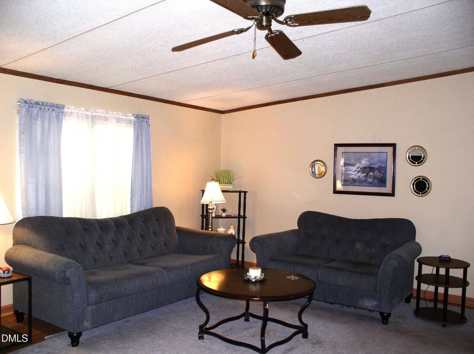 A cozy living room with two gray sofas, a round wooden coffee table, a side table with a lamp, and a window with light blue curtains. A ceiling fan and framed wall art are also visible.