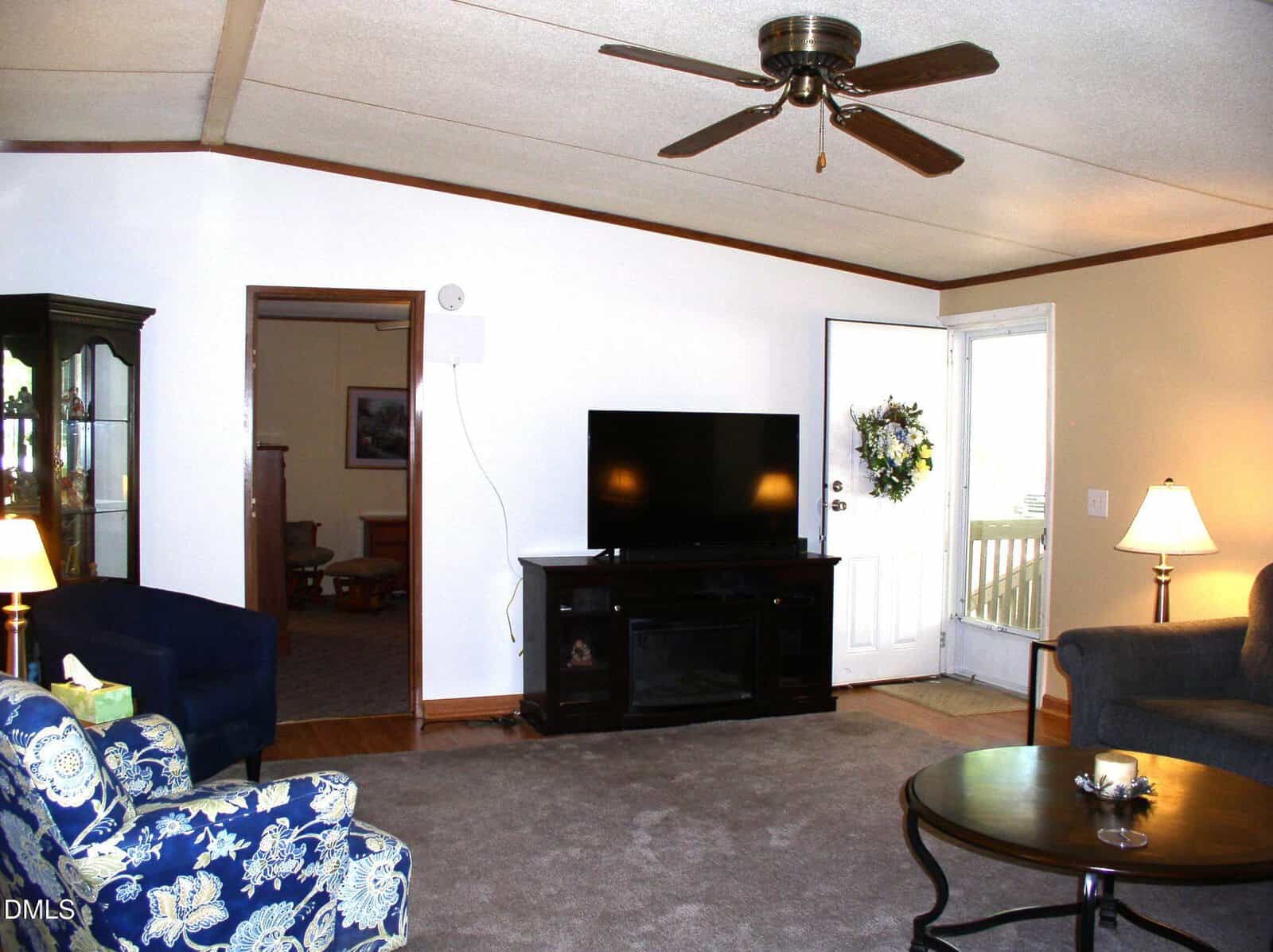 A living room with beige carpet, a TV on a black stand, a round wooden coffee table, blue and floral armchairs, a gray sofa, a lamp, and a ceiling fan. An open door with a wreath leads to a porch.