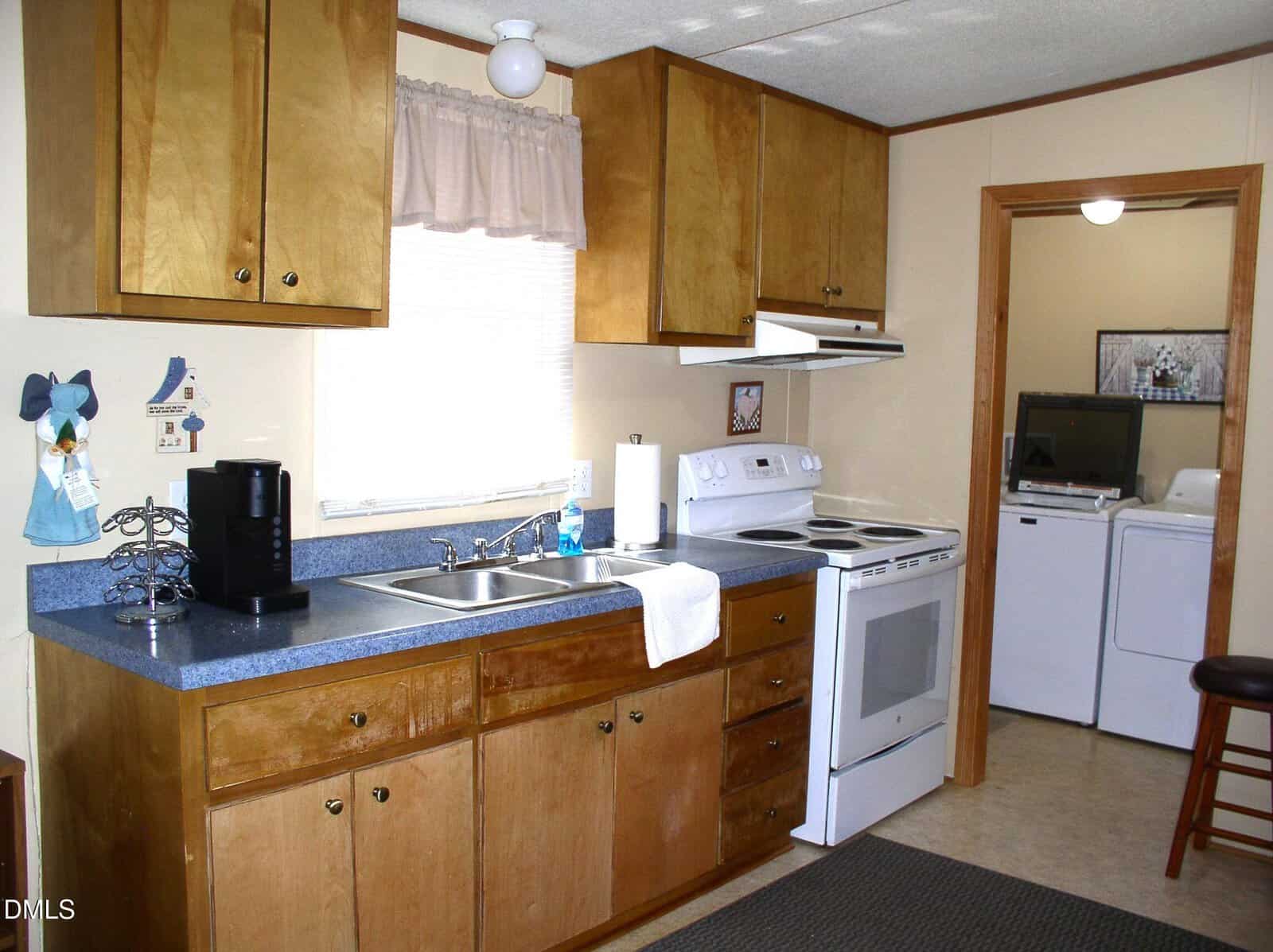 A kitchen with wooden cabinets, blue countertops, a white stove and oven, a coffee maker, a double sink, and a towel hanging on the counter. A laundry room with a washer and dryer is visible through a doorway.