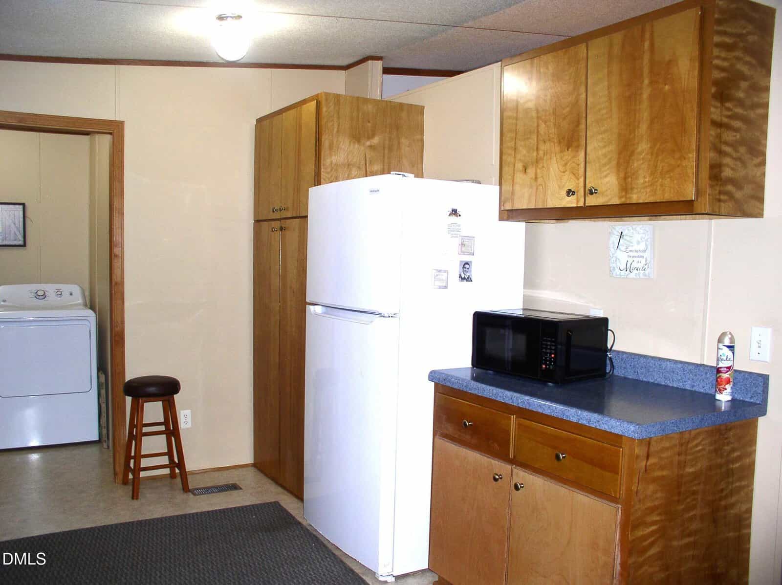 A kitchen with wooden cabinets, a white refrigerator, a microwave on a blue countertop, and a can of air freshener. In the background, there is a laundry area with a washer, dryer, and a wooden stool.